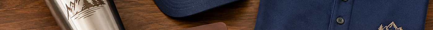 Branded company event merch on a wood table: engraved tumbler, leather patch hat, embroidered polo, leatherette journal, and acacia coasters
