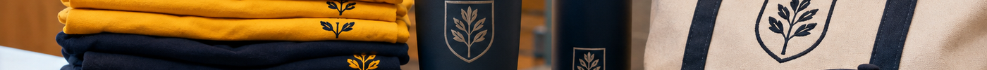 School spirit wear on a table in a gym lobby - stacked navy and gold tees, a grey crewneck sweatshirt, an engraved tumbler, an insulated water bottle, a canvas tote, and a leather patch cap